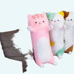 On a grey sofa in a small living room with a brown floor, 3 plush pillows with the effigy of a big cat standing on the sofa, one is pink, the second is green and the third is brown