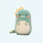 Soft Squishmallow plush shaped like a green dinosaur with a beige belly, pink cheeks, and a sweet smile. It features cute details like a yellow crest on its head and a yellow-spiked tail, making it extra cuddly and lovable.